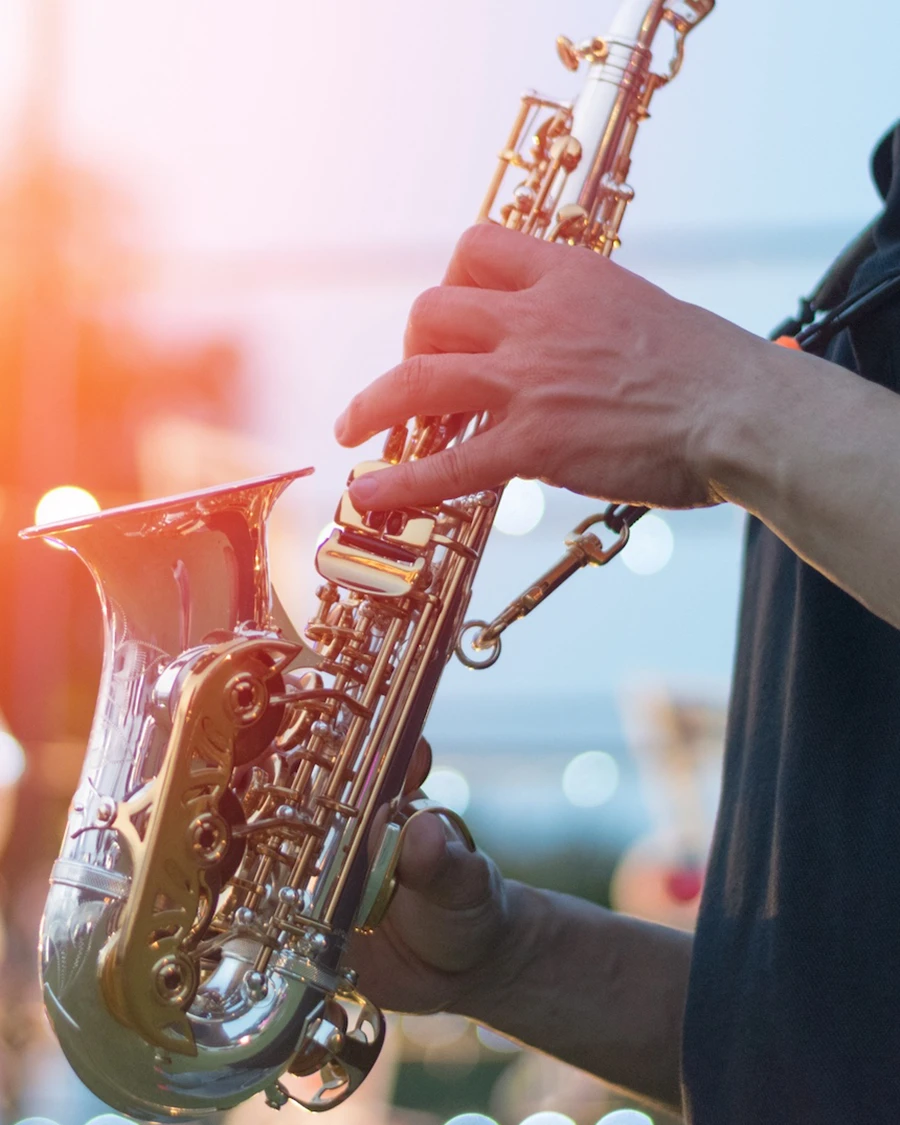 Close-up of hands playing a silver and gold saxophone outdoors at sunset, with festival lights glowing in the background